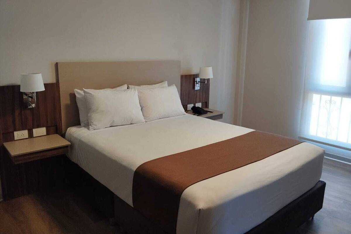 A hotel room with a large bed, two bedside tables, and a window with blinds.