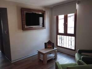 Una sala de estar con un televisor de pantalla plana montado en la pared, una pequeña mesa de madera y un sillón verde.