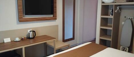 A hotel room with a bed, a desk with a kettle, a mirror, a wardrobe, and a television.