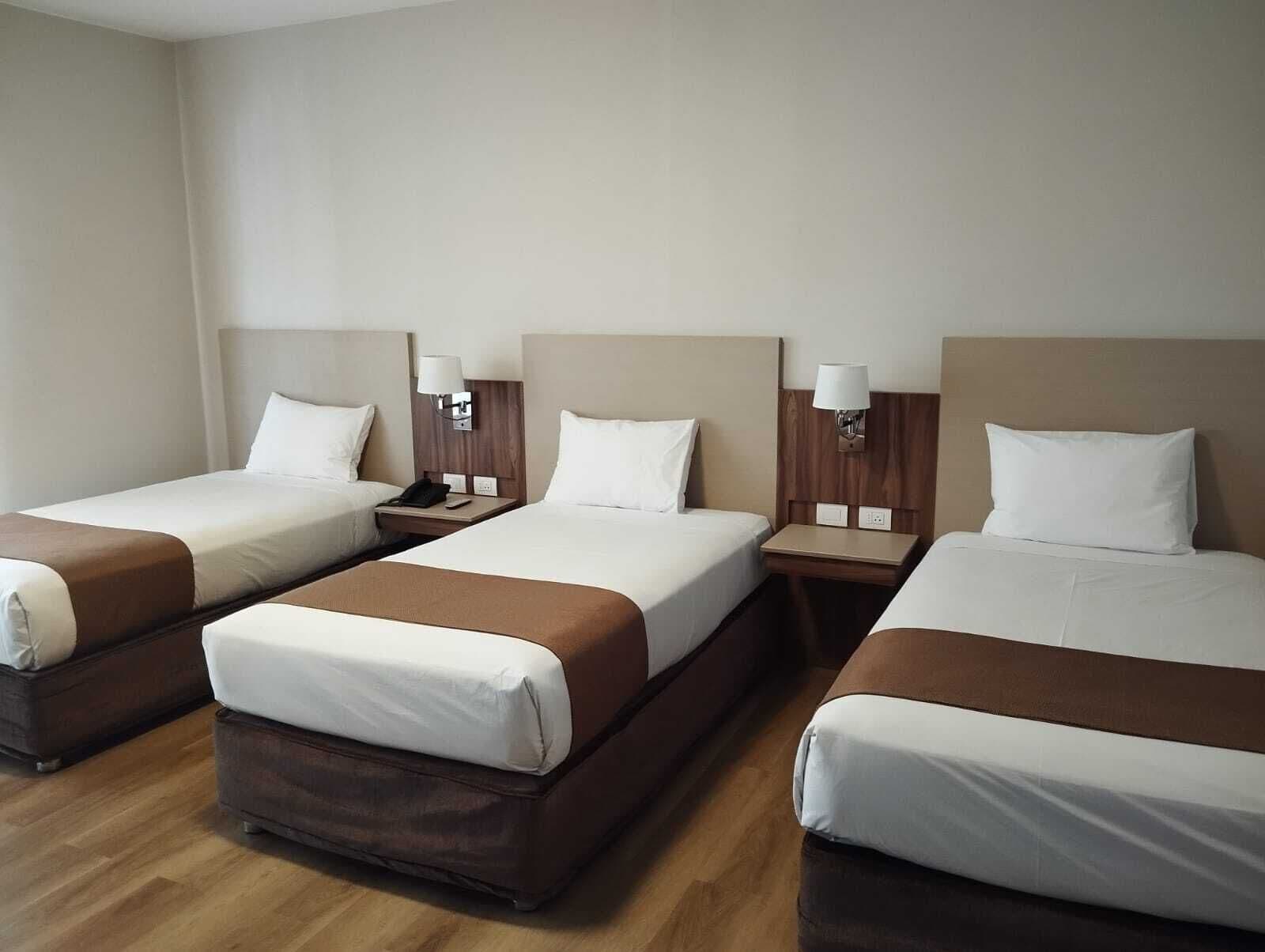 Habitación de hotel con tres camas individuales, cada una con cabecera de madera, ropa de cama blanca y una mesita de noche con lámpara.