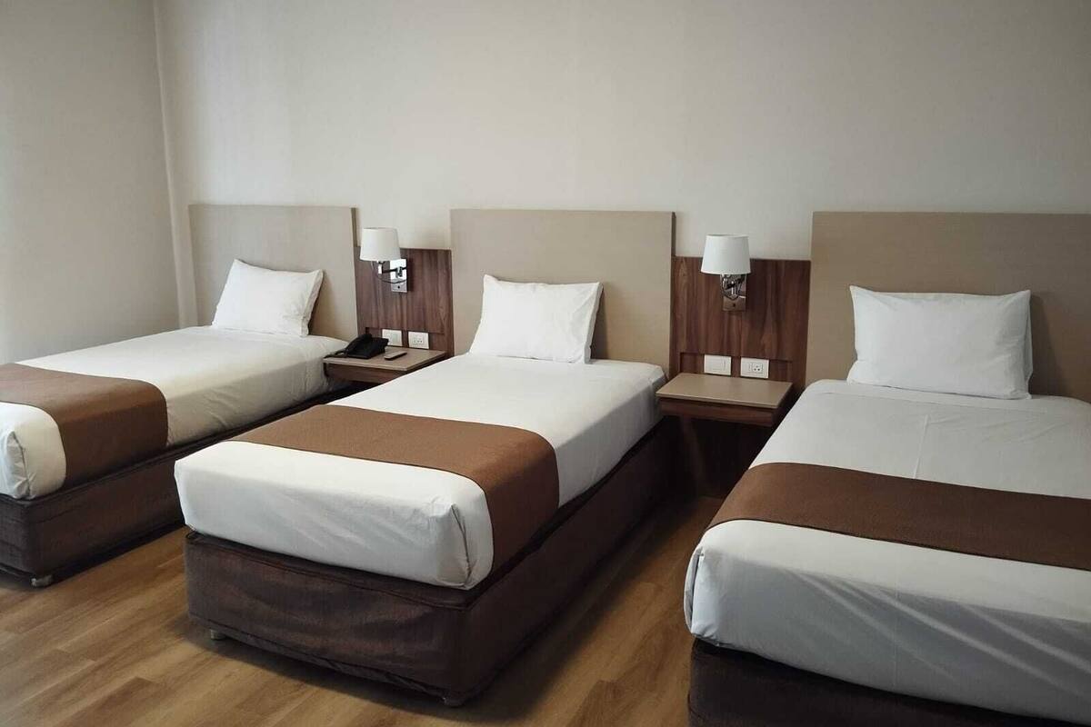 A hotel room with three single beds, each with a wooden headboard, white bedding, and a bedside table with a lamp.