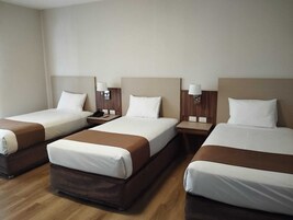 Habitación de hotel con tres camas individuales, cada una con cabecera de madera, ropa de cama blanca y una mesita de noche con lámpara.