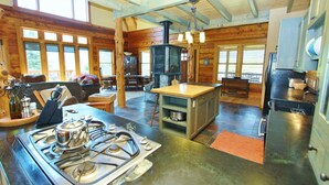 A rustic cabin interior with a kitchen area featuring a gas stove, wooden dining table, and a stone fireplace. - Ashling Ranch - Gorgeous, luxury log home with access to Yellowstone River! (Emigrant)