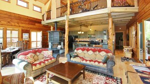A spacious log cabin interior with a high ceiling, a central fireplace, and a kitchen area. - Ashling Ranch - Gorgeous, luxury log home with access to Yellowstone River! (Emigrant)
