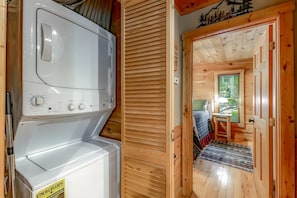 A laundry area with a front-loading washer and dryer, a wooden door leading to a bedroom with a bed and a window, and a small table with a lamp.