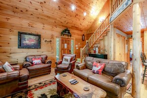 A log cabin interior with leather sofas, a coffee table, and a staircase leading to a loft area.