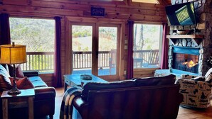 Ein gemütliches Cabin-Interieur mit Holzbalkendecke, Kamin, Fernseher an einer Steinwand und einem großen Fenster mit Blick auf eine Terrasse.