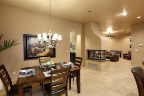 A dining area with a wooden table, chairs, and a chandelier. There is a painting on the wall and a fireplace in the background.