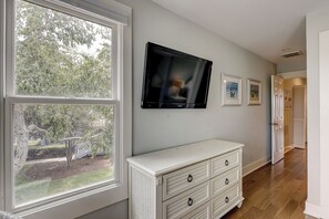 Una habitación con un ventanal grande, un televisor de pantalla plana montado en la pared, un aparador blanco con cajones y una puerta que da a otra estancia.