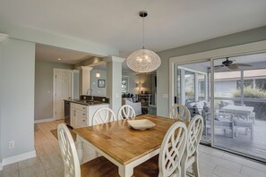 Un coin repas avec une table en bois et des chaises blanches, une cuisine équipée d’un évier et de placards, et une grande porte-fenêtre coulissante donnant sur un patio extérieur.