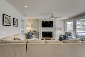 A living room with a sectional sofa, a fireplace, a ceiling fan, and a TV mounted on the wall. - Beautiful Lands End Townhome with Spectacular Water Views & Close to Beach (Hilton Head Island)