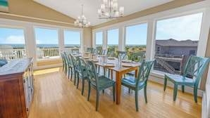 A dining area with a view of the ocean, featuring a wooden table set for six, teal chairs, and a chandelier. - Premier Dog-Friendly Oceanfront 9BR w/ Private Pool, Tiki Bar, Theater, Elevator (Salvo)