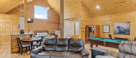 Un intérieur de chalet chaleureux avec un coin repas, un canapé, une table de billard et une grande fenêtre.