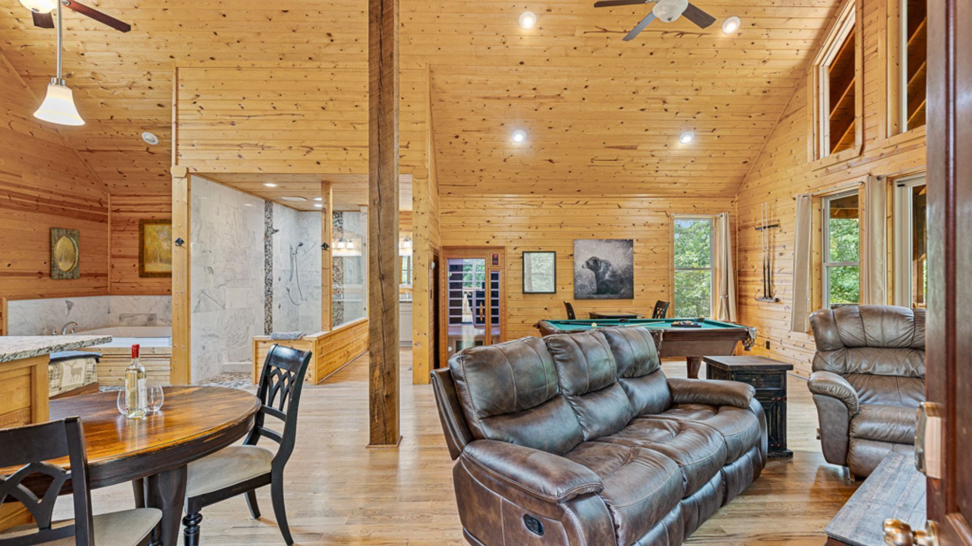A spacious room with wooden walls and ceiling, a pool table, leather sofas, and a dining area with a table and chairs.