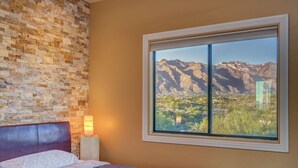 A bedroom with a brick wall, a large window with a mountain view, a bed with a headboard, and a nightstand with a lamp.