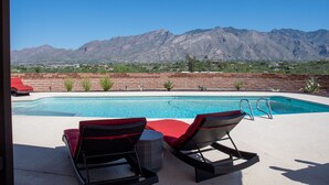 A pool with two wicker chairs and a red cushion, surrounded by a brick wall and a view of mountains.
