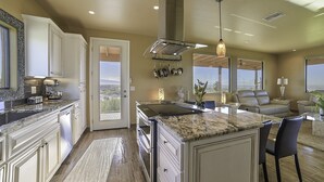 A modern kitchen with granite countertops, white cabinetry, and a large island. There is a dining area with a leather couch and a view of the outdoors.