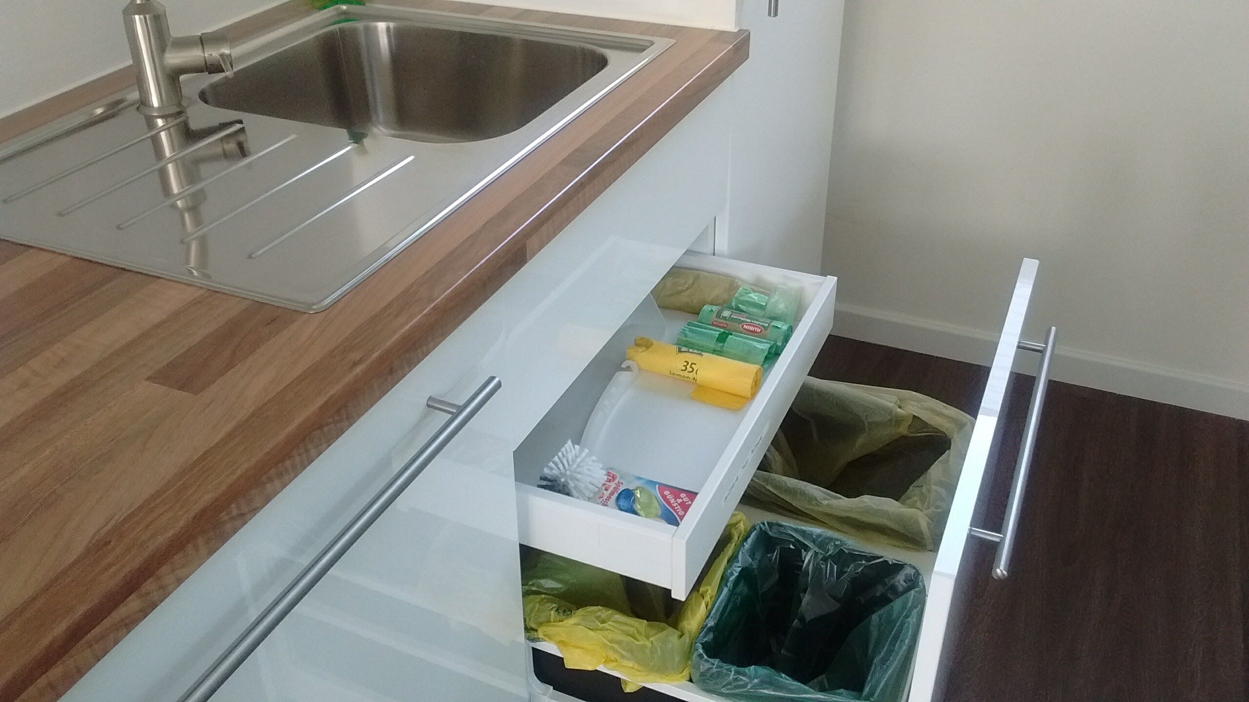 A kitchen with a stainless steel sink, a wooden countertop, and a white drawer with trash bags and cleaning supplies.