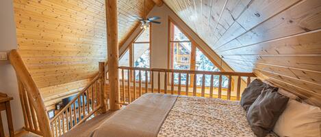 Un dormitorio con techo inclinado, vigas de madera, una cama con ropa de cama estampada y vistas a una casa a través de ventanas grandes.