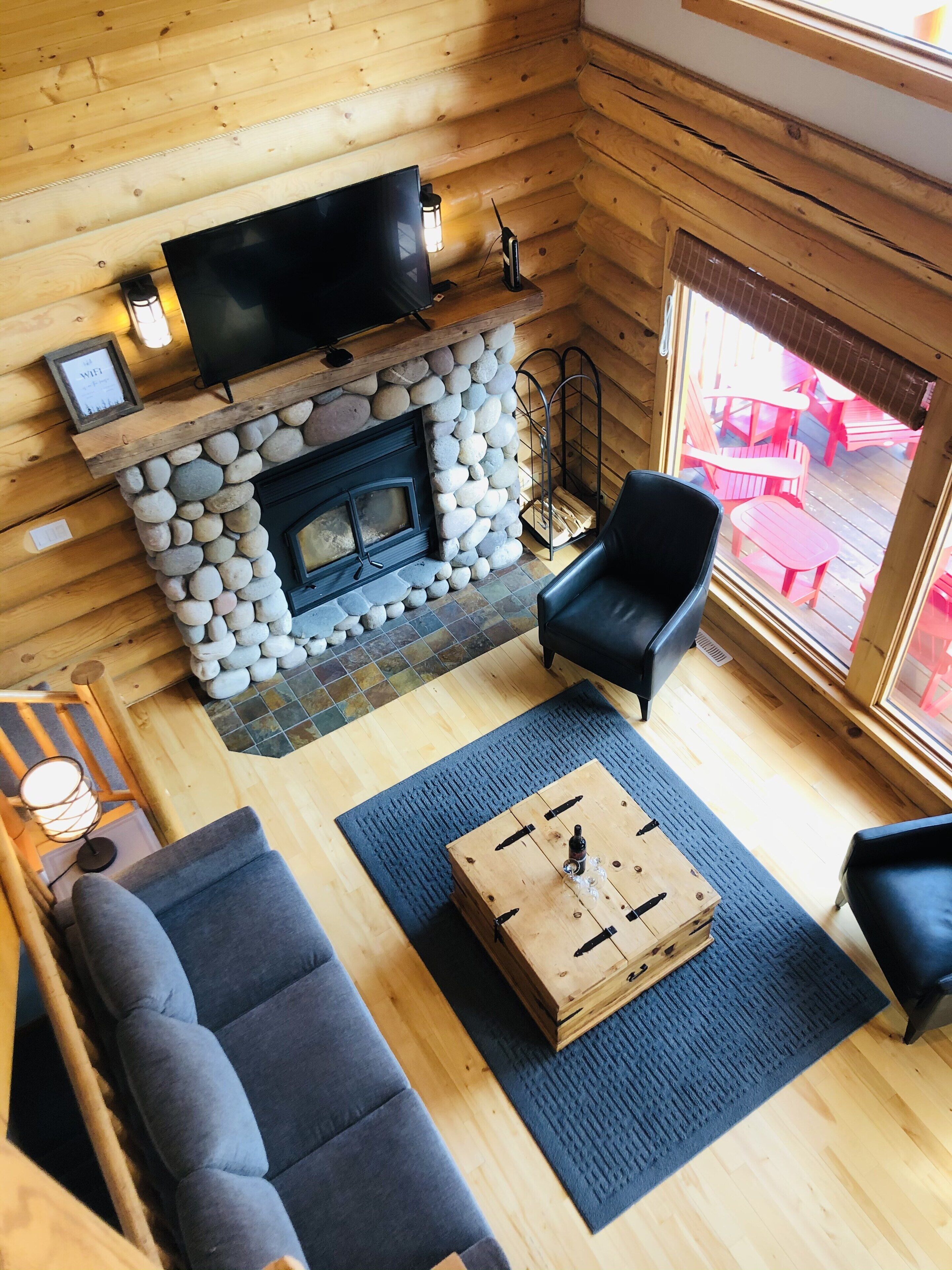 Ein Interieur einer Blockhütte mit Kamin aus Stein, einem Flachbildfernseher, einer Couch und einem Essbereich mit roten Stühlen.