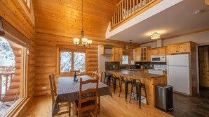 Una cabaña de troncos con interior de madera, un comedor con mesa y sillas, una cocina con electrodomésticos modernos y un balcón con barandilla de madera.