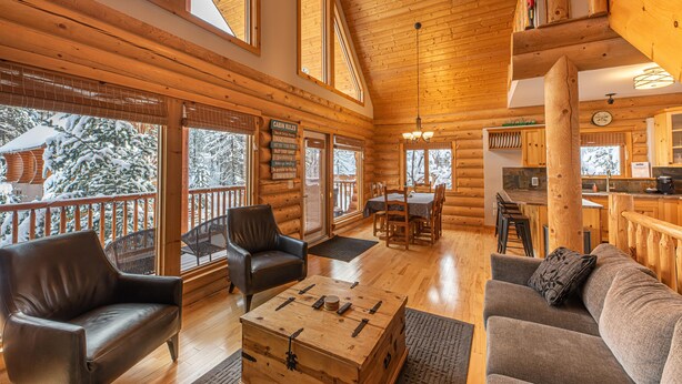 Interior de una cabaña de troncos con una sala de estar acogedora, muebles de madera y grandes ventanas que ofrecen vistas a árboles cubiertos de nieve.