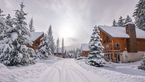 Una cabaña nevada con fachada de madera, una entrada visible y un camino de acceso a la edificación principal.