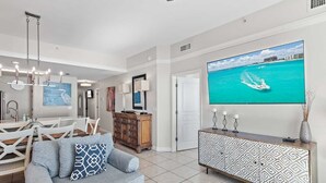 Una sala moderna con un televisor de pantalla plana grande mostrando una playa tropical, un comedor con una mesa y sillas blancas, y un aparador con motivos decorativos.