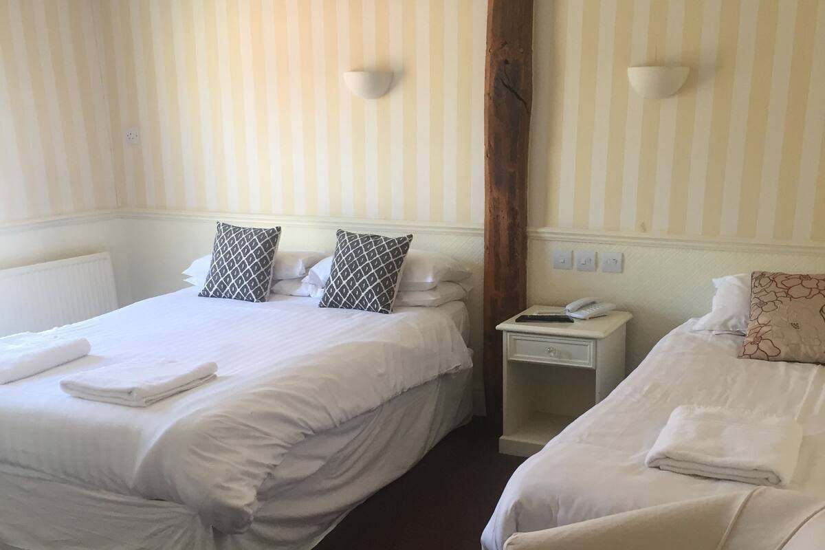 A hotel room with two beds, striped wallpaper, a wooden beam, a nightstand with a phone, and a radiator.