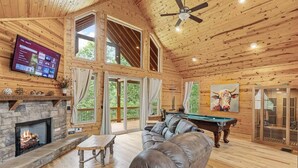 Ein gemütliches Blockhaus-Interieur mit Kamin aus Stein, Ledersofa, Billardtisch und einem an der Wand montierten Fernseher.