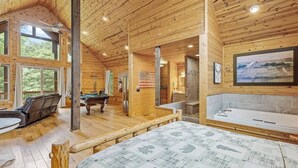 Ein Holzblockhaus-Interieur mit Billardtisch, einer Couch, einer Badewanne und einem großen Fenster mit Blick ins Freie.