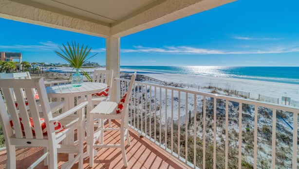 A balcony with white chairs and a table, overlooking a beach with a fence and a palm tree.