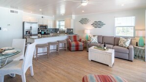 A modern kitchen with white cabinets, a stainless steel refrigerator, and a microwave. There is a dining area with white wicker chairs and a glass table. A living room with a grey sofa and colorful cushions is visible.