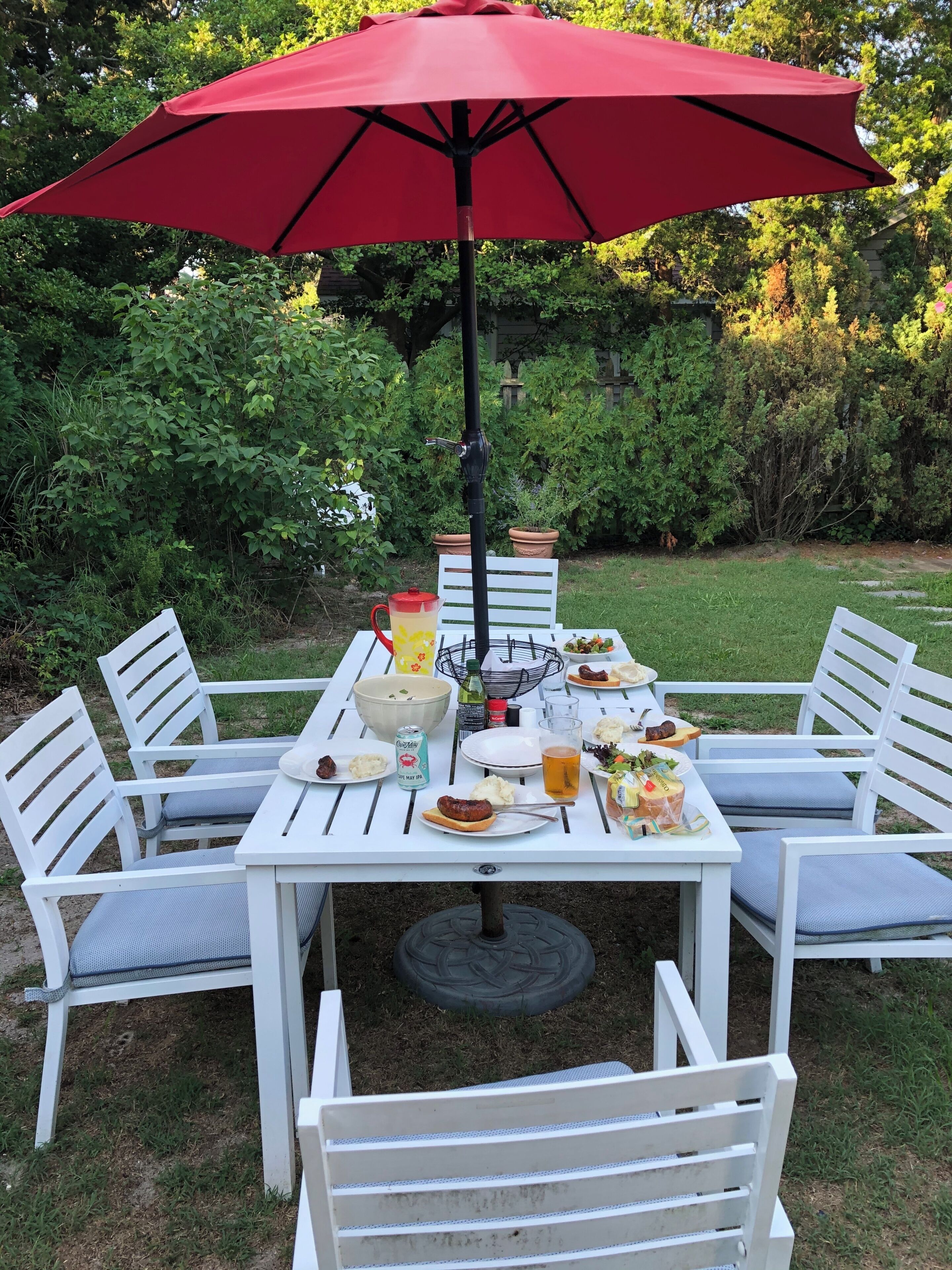Mesa blanca al aire libre con una sombrilla roja, rodeada de sillas blancas, ubicada en un jardín con follaje de fondo.