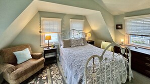 Une chambre avec un plafond en pente, un lit avec une tête de lit blanche, une table de chevet avec une lampe, une fenêtre avec des stores et une vue sur une clôture blanche à l’extérieur.