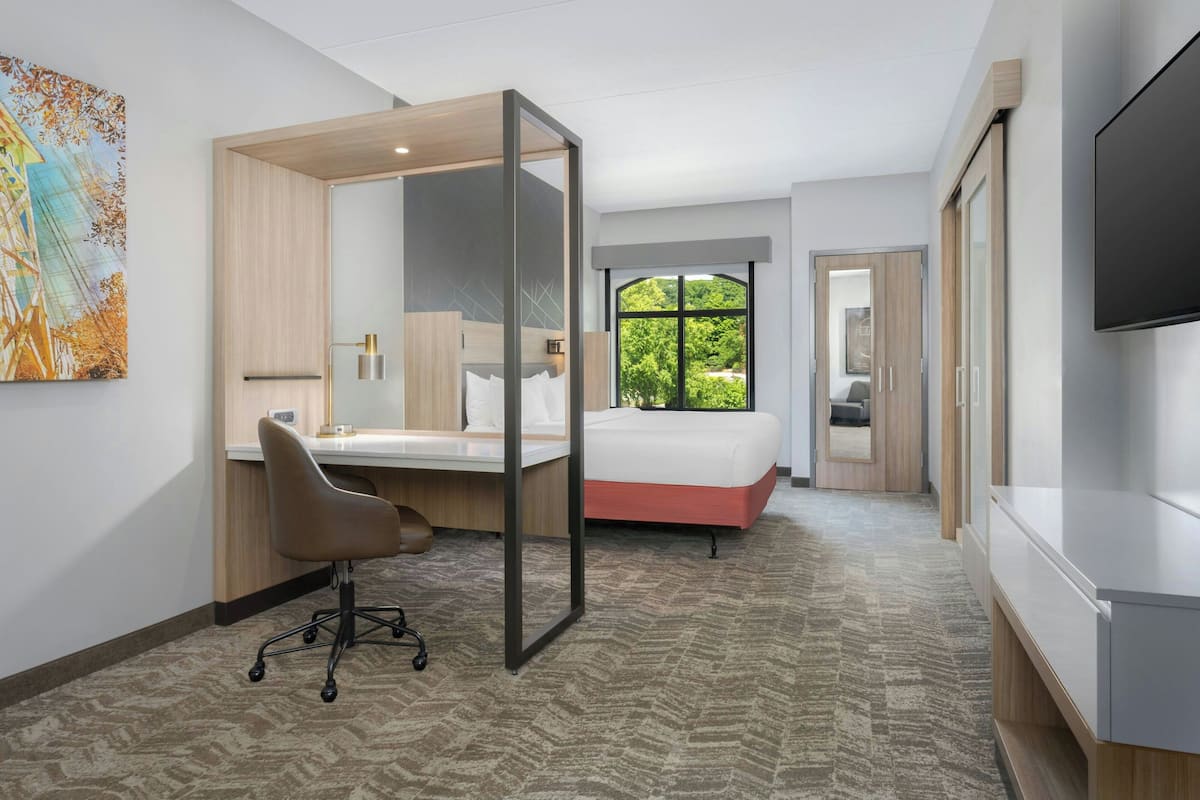 A modern hotel room with a bed, desk, chair, and a large window offering a view of greenery.