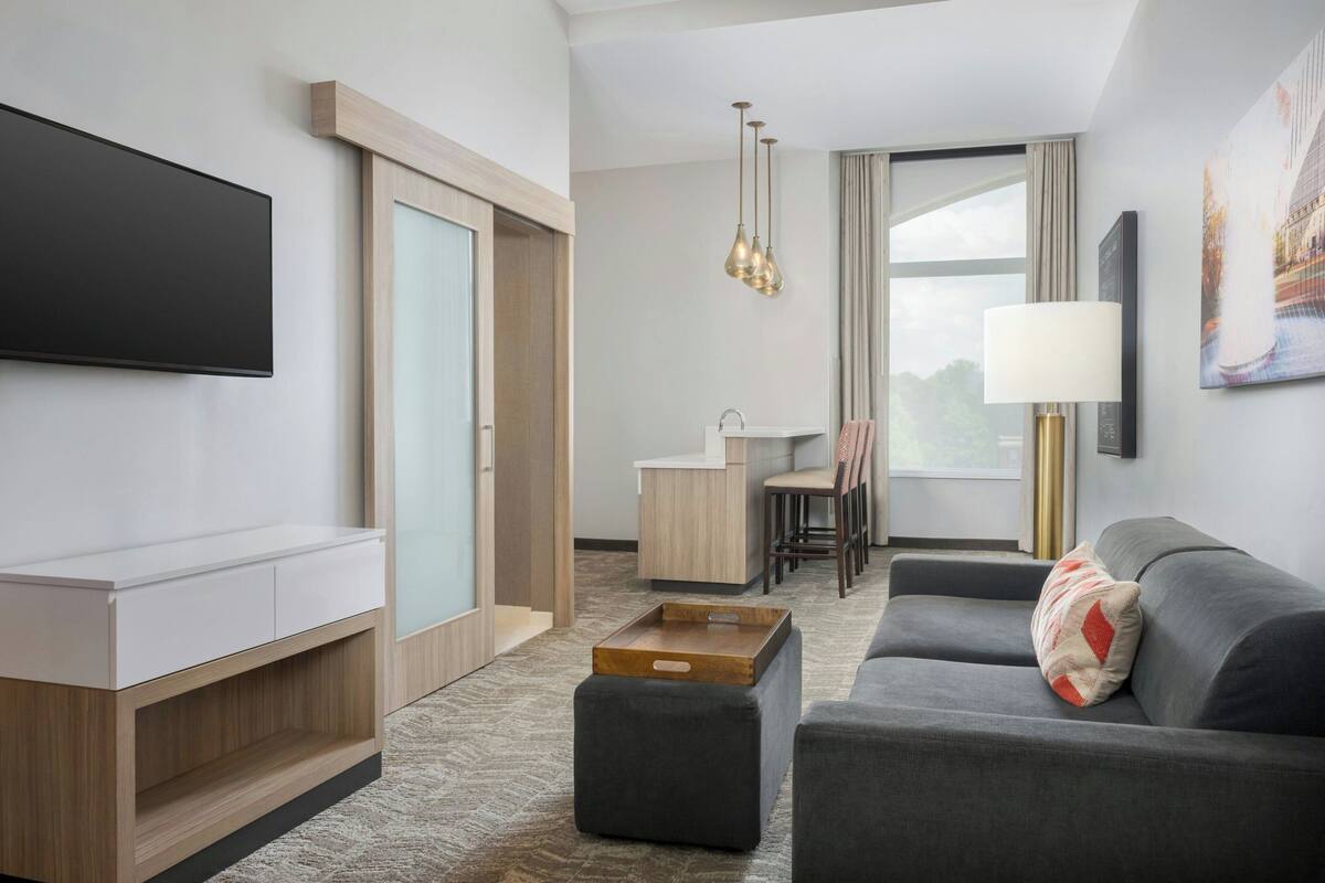 A modern hotel room with a flat-screen TV, a sofa, a coffee table, a desk with a chair, and a large window.