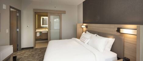 A hotel room with a large bed, a bedside table, a nightstand, a mirror, and a bathroom visible through an open door.
