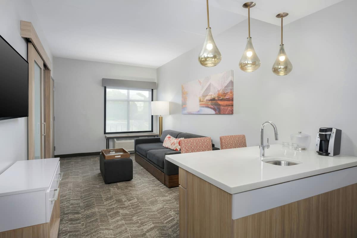 A modern kitchen with a white countertop, stainless steel sink, and a coffee machine. There is a gray sofa with patterned cushions and a window providing natural light.