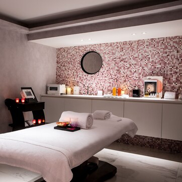 A modern spa room with a massage table, a small side table with candles, and a reception area with a tiled wall and various toiletries.