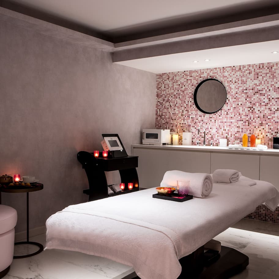 A modern spa room with a massage table, a small side table with candles, and a reception area with a tiled wall and various toiletries.