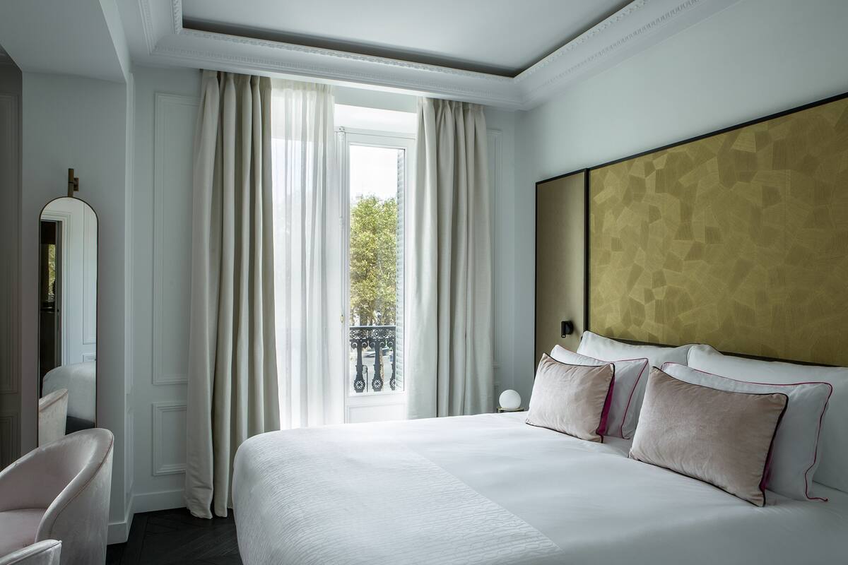 A hotel room with a large bed, a nightstand, a chair, and a view of the outdoors through a window with curtains.