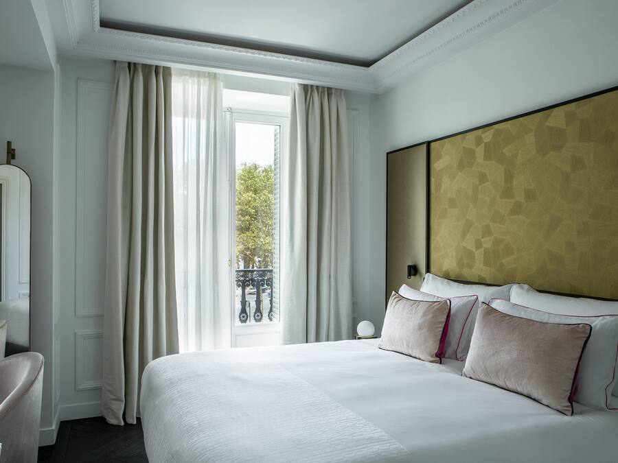 A hotel room with a large bed, a nightstand, a chair, and a view of the outdoors through a window with curtains.