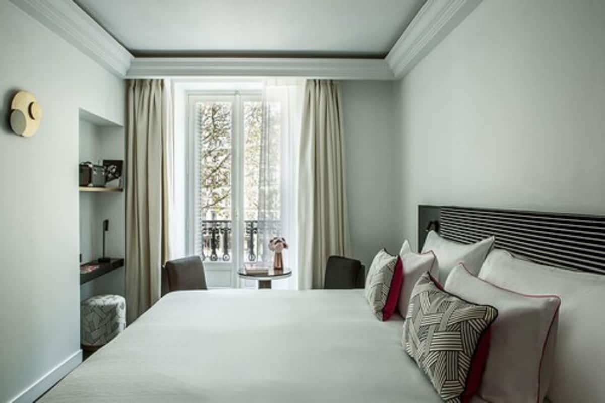 A hotel room with a large bed, a small table with a vase, a chair, and a shelf with a camera.
