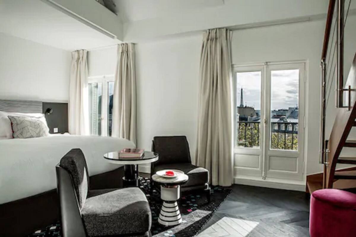 A hotel room with a bed, a small round table, two chairs, a red ottoman, and a view of the city through large windows.