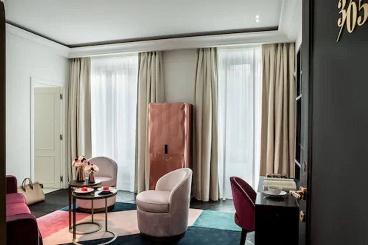 A modern hotel room with a large window, a pink chair, a small round table, and a wardrobe.