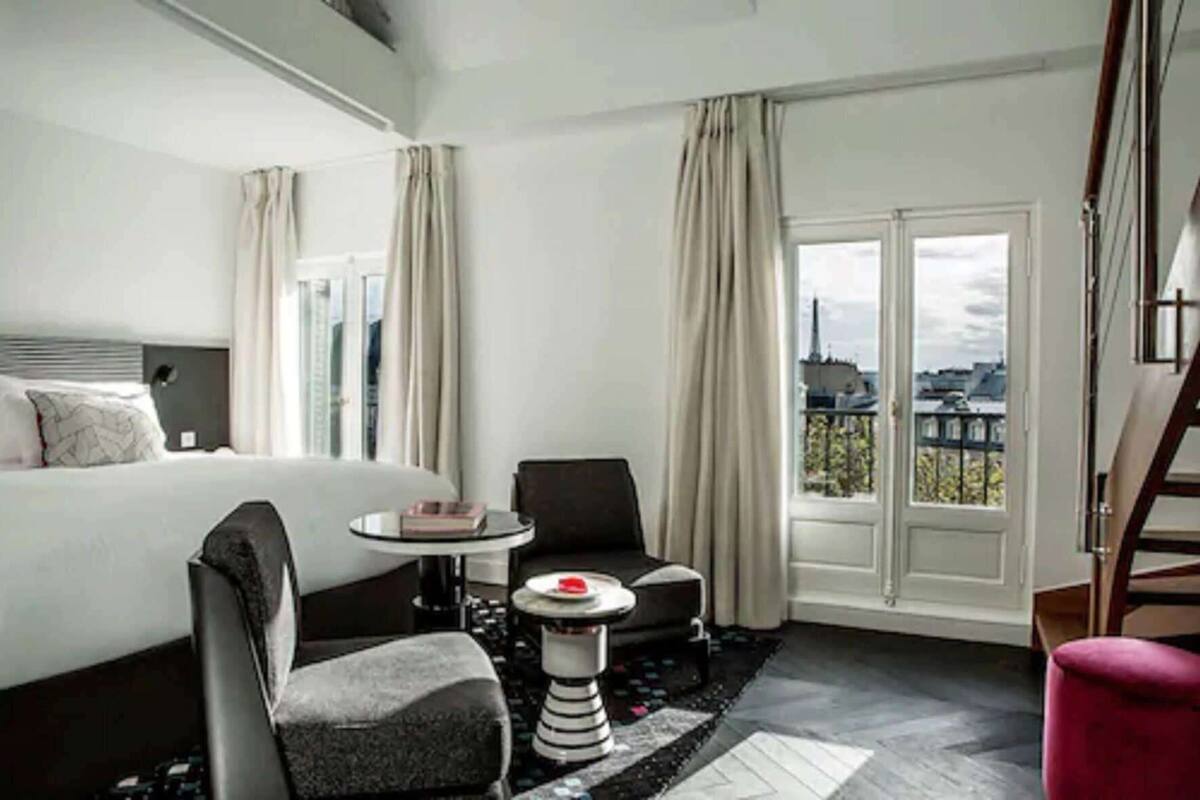 A hotel room with a bed, a small round table, a chair, a red ottoman, and a view of buildings through a large window.