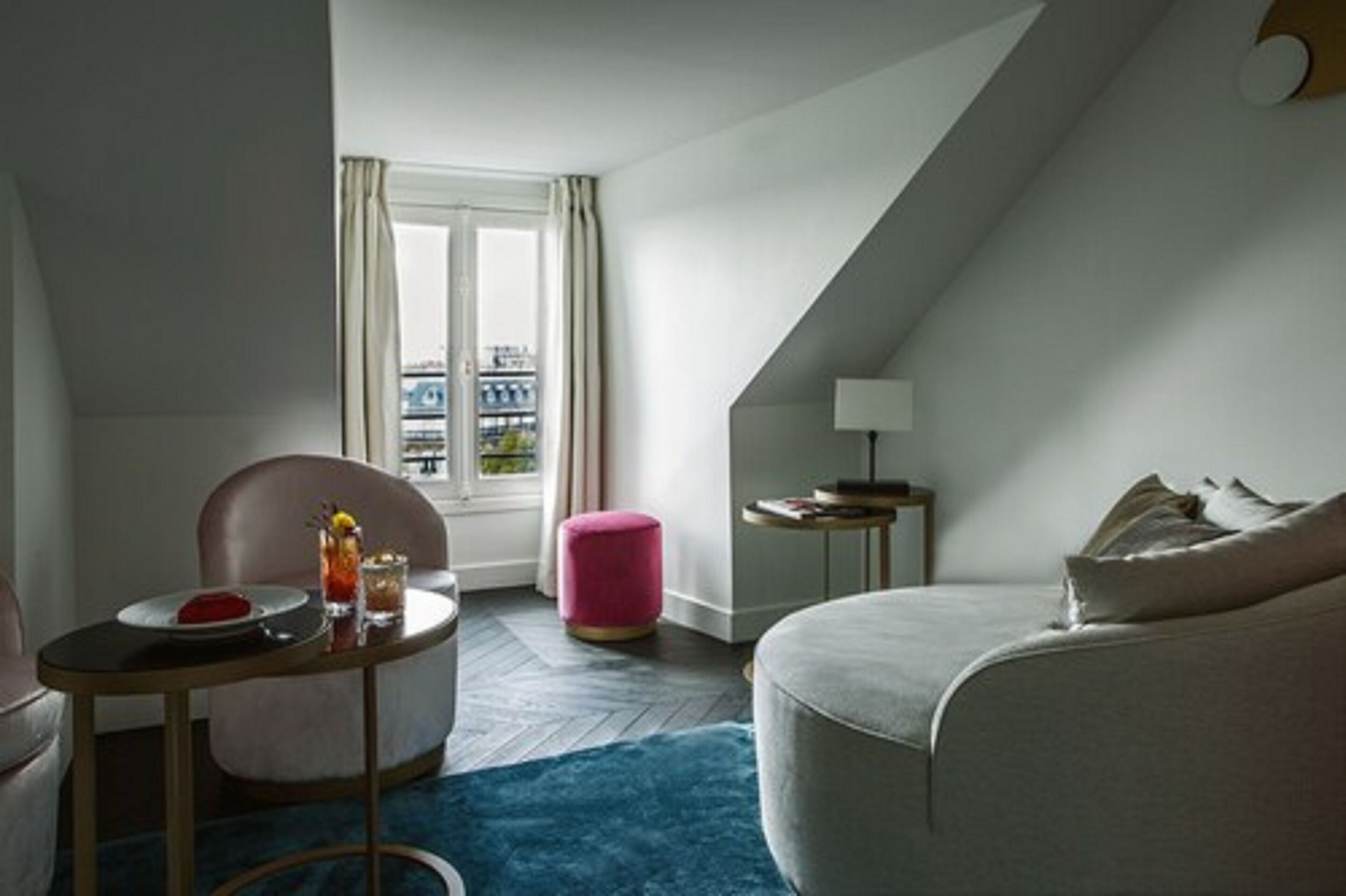 A modern hotel room with a cozy seating area, a small table with a drink, a window with curtains, and a lamp on a side table.