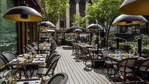 Un área de comedor al aire libre con mesas y sillas, sombrillas grandes y vista a un edificio con columnas.