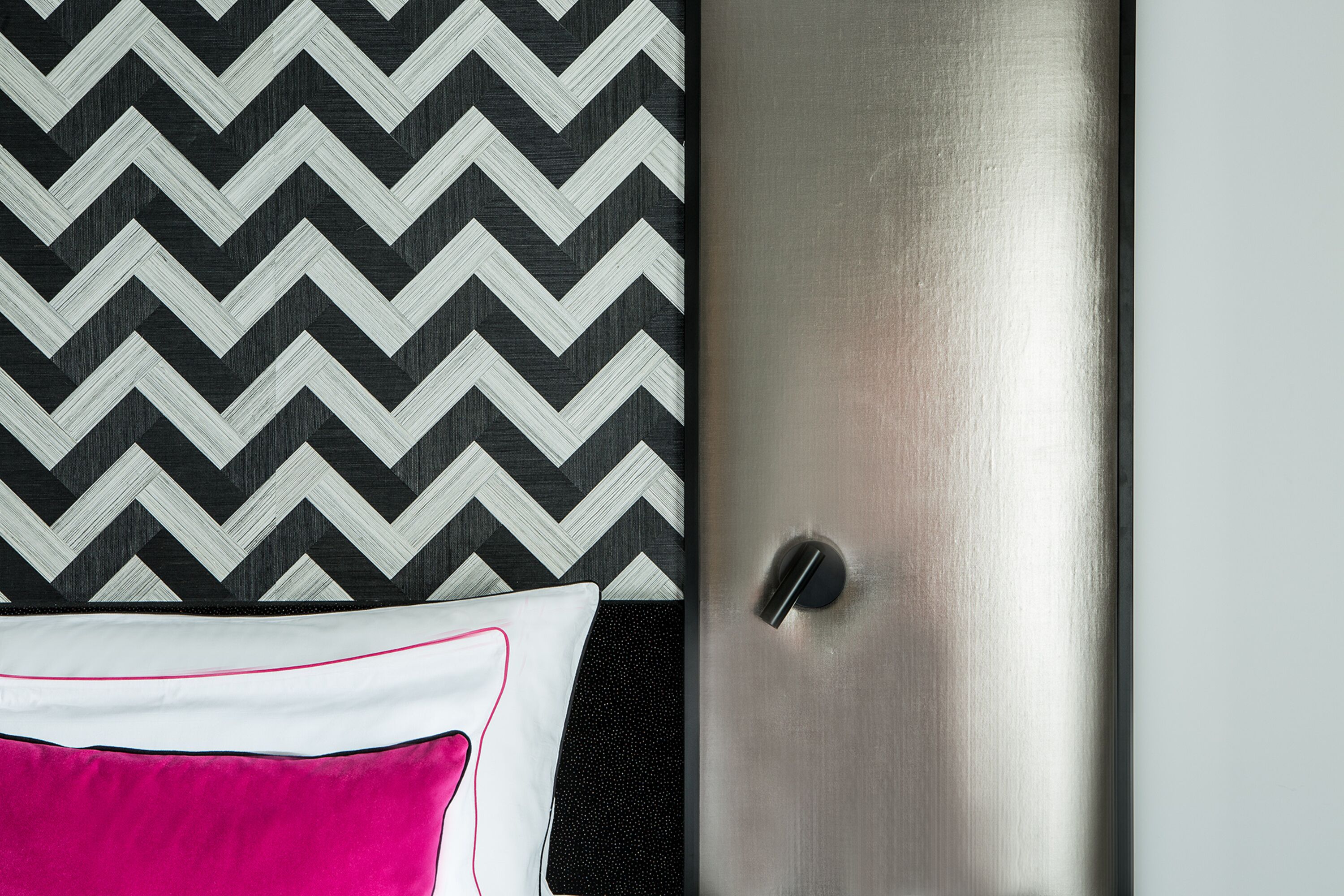 A hotel room with a metallic door, a black and white chevron-patterned wall, and a white pillow with pink piping.